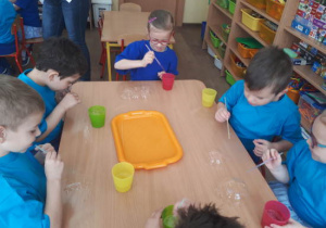 Dzieci przy stole próbują posadzić bańkę mydlaną na blacie stolika.